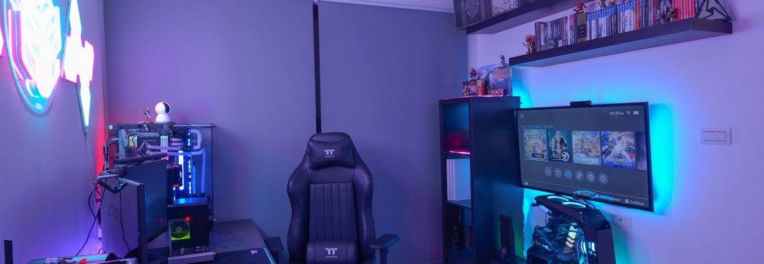A modern gaming setup featuring a sleek gaming chair, dual monitors on a desk, and LED lighting. A gaming console and various decorations are displayed on shelves, creating an organized and vibrant gaming environment.