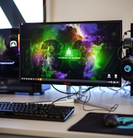 A gaming setup featuring a central monitor with a vibrant display showing 'The Spirit of Gaming'. On either side, there are gaming PCs with illuminated components and transparent cases. Headsets hang from the side of the monitor. The desk also has a black keyboard and mouse, and a branded mousepad.