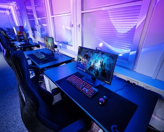 A row of modern gaming setups is arranged neatly along a window-lined wall. Each station includes a high-resolution monitor displaying vivid graphics, a mechanical keyboard with colored lighting, a gaming mouse, and an ergonomic chair. The room is illuminated with ambient blue and purple lights, creating a sleek and futuristic atmosphere.