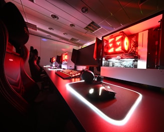A modern gaming setup featuring several high-end computers with illuminated red interior lighting. The room is equipped with gaming chairs exhibiting a sleek black and red design. Multiple monitors are arranged on a long desk, and peripheral devices like keyboards and mice are uniformly placed, reflecting an ambiance ideal for gaming or competitive play.
