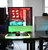 A modern gaming setup is displayed on a desk with multiple computer monitors and an open gaming PC tower. The tower is illuminated with red and green LED lights, showcasing a 'GeForce RTX' graphics card. A gaming mouse rests on a mousepad with glowing edges, and there is a keyboard with vibrant backlighting. The room appears well-lit with visible windows in the background.