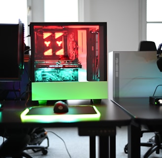 A modern gaming setup is displayed on a desk with multiple computer monitors and an open gaming PC tower. The tower is illuminated with red and green LED lights, showcasing a 'GeForce RTX' graphics card. A gaming mouse rests on a mousepad with glowing edges, and there is a keyboard with vibrant backlighting. The room appears well-lit with visible windows in the background.