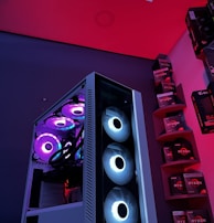 A gaming computer setup featuring a high-performance PC tower with multiple RGB fans lit in blue and purple hues. The case has a modern design with transparent panels showcasing internal components. In the background, shelves are stocked with various Ryzen and other gaming components under a vibrant red and blue ambient light.