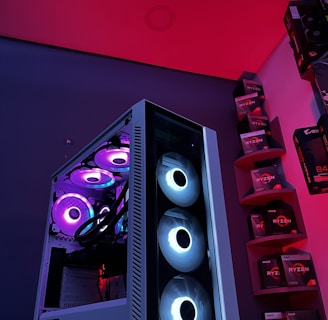 A gaming computer setup featuring a high-performance PC tower with multiple RGB fans lit in blue and purple hues. The case has a modern design with transparent panels showcasing internal components. In the background, shelves are stocked with various Ryzen and other gaming components under a vibrant red and blue ambient light.