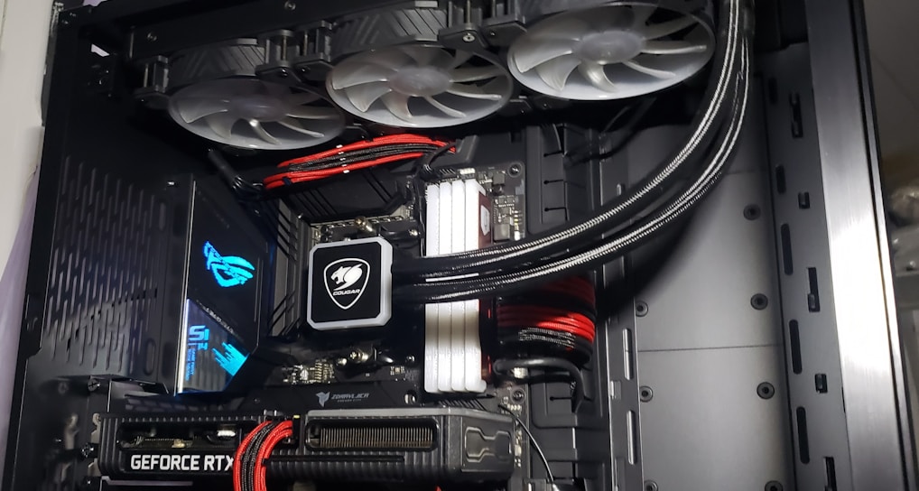 A high-performance gaming computer setup showing the interior components, including a GeForce RTX graphics card, cooling system with three fans, a motherboard, and visible wiring with red and black cables.