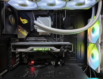 Inside view of a computer case showcasing a sophisticated gaming setup. Visible components include a GeForce RTX graphics card illuminated with green lighting, an ASUS motherboard, cooling fans with LED lighting, and a liquid cooling system with white tubes. Cables are neatly organized, and overall, the hardware suggests a high-performance machine.