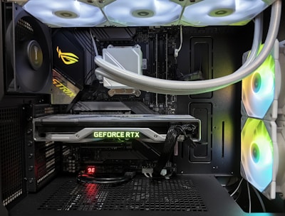Inside view of a computer case showcasing a sophisticated gaming setup. Visible components include a GeForce RTX graphics card illuminated with green lighting, an ASUS motherboard, cooling fans with LED lighting, and a liquid cooling system with white tubes. Cables are neatly organized, and overall, the hardware suggests a high-performance machine.